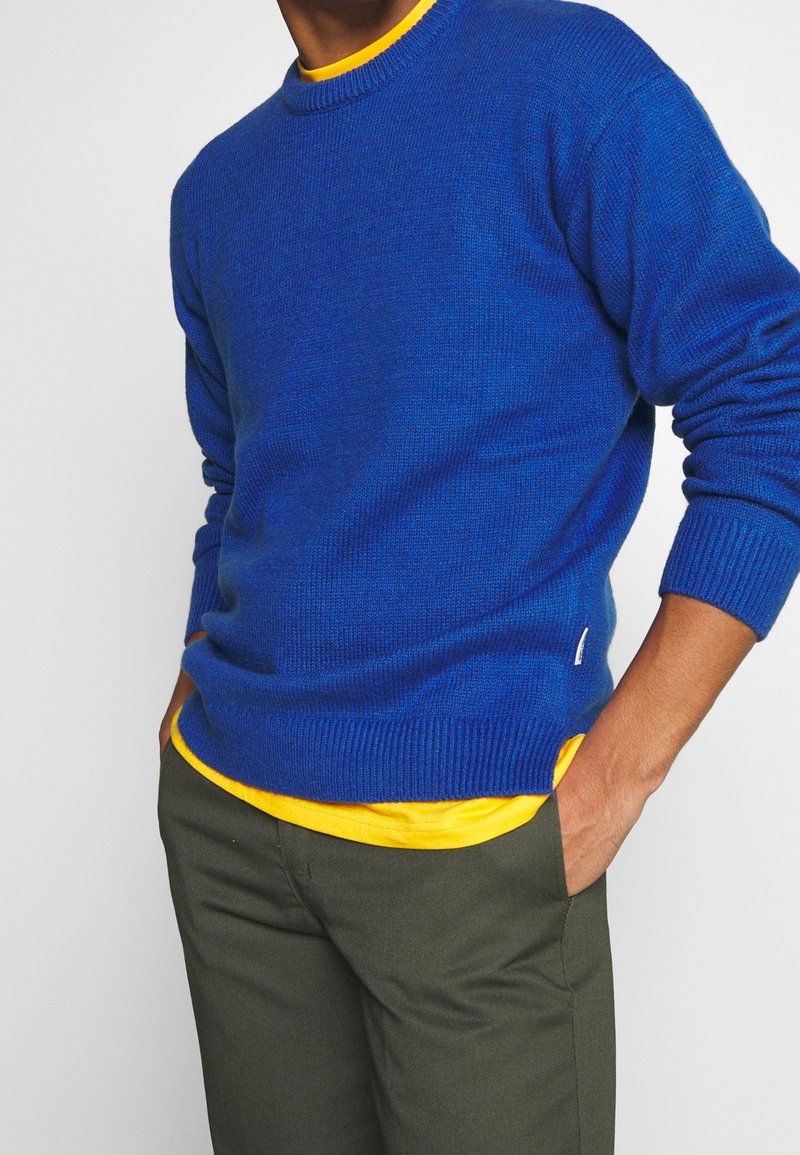 Niebieski sweter z dzianiny z okrągłym dekoltem, z prążkowanymi mankietami i dołem, nałożony na żółtą koszulkę, w zestawieniu z oliwkowymi spodniami.