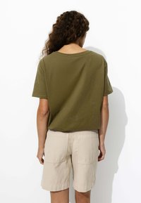 T-shirt en coton vert olive à coupe courte et décontractée, associé à un short beige jusqu'aux genoux avec deux poches arrière. Fond blanc.