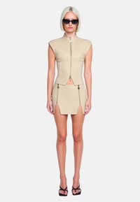 Top corto beige con colletto alto e chiusura a zip abbinato a una mini gonna coordinata con spacchi e zip. Occhiali da sole neri e scarpe.