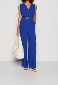 Blå ärmlös jumpsuit med V-ringning, vida ben och ett bälte med guldspänne. Matchas med en krämfärgad väska och vita sandaletter med blockklack.