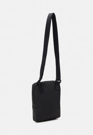 Sac bandoulière rectangulaire noir avec une longue bandoulière réglable et une fermeture éclair sur le dessus, sur un fond uni clair.