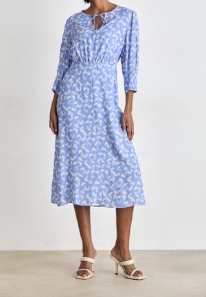 Žena ve světle modrých midi šatech s bílými abstraktními vzory, tříčtvrtečními rukávy, s vázačkou na krku a béžovými sandály na vysokém podpatku.