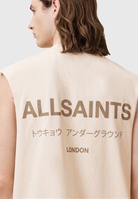 Beige ermeløs skjorte med løs passform, med stor brun tekst: "ALLSAINTS" og "LONDON" på baksiden. Myk bomullsstruktur.