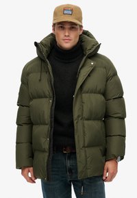 Olivgrüne Puffjacke mit Kapuze, ausgestattet mit einem Reißverschluss vorne, großen gesteppten Bereichen und Kordelzügen. Über einem schwarzen Rollkragenpullover getragen.