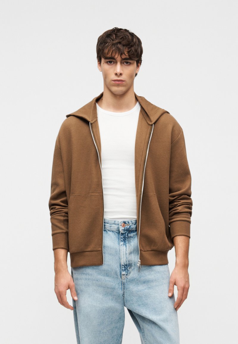 Καφέ φούτερ με φερμουάρ και ρτιγμένη υφή, μεγάλο κουκούλι και εμπρόσθιες τσέπες, συνδυασμένο με ανοιχτό μπλε τζιν παντελόνι χαλαρής γραμμής.