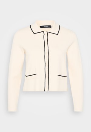 Cárdigan crema con bordes negros, que presenta un cuello, botones frontales y dos bolsillos inferiores con detalles en negro. Textura de punto suave.