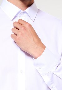 Camicia bianca con colletto classico, chiusura frontale con bottoni e polsini. Texture liscia, design aderente e dettagli di cucitura resistenti.