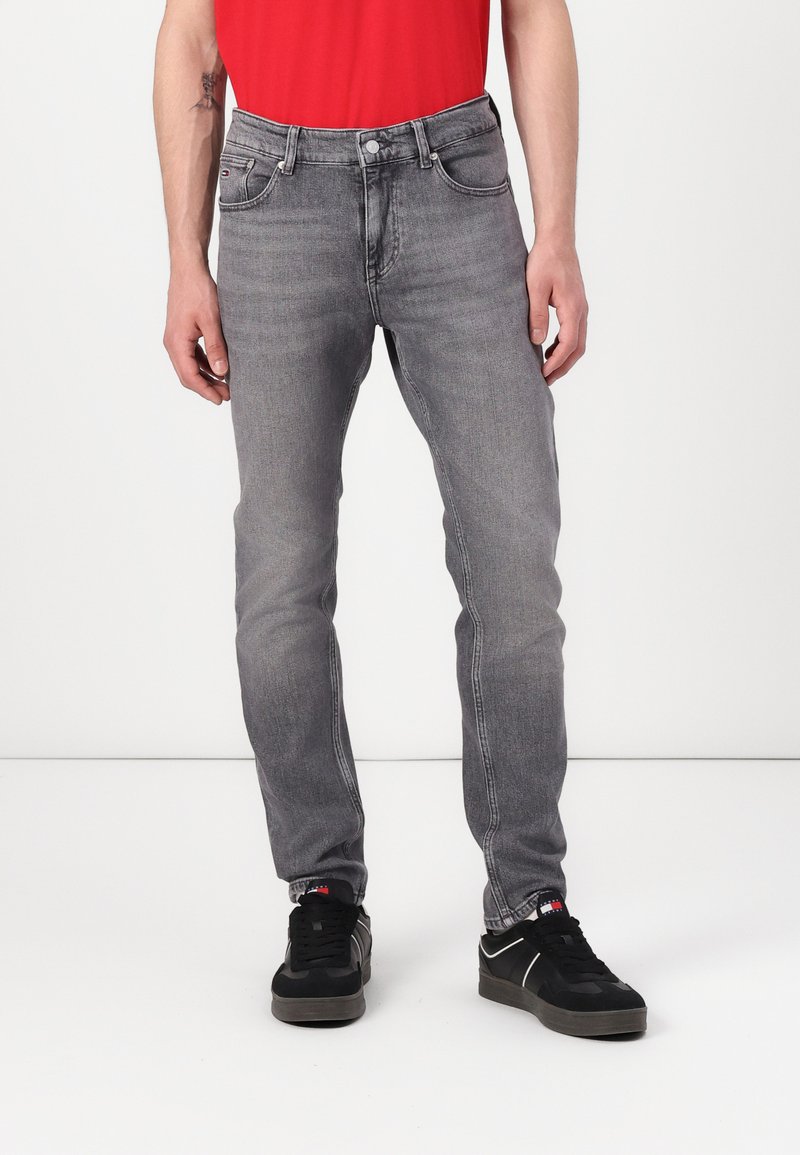 Graue Denim-Jeans mit schlankem Schnitt, Fünf-Taschen-Design und subtiler Waschung. Kombiniert mit schwarzen Sneakers und einem roten Hemd.