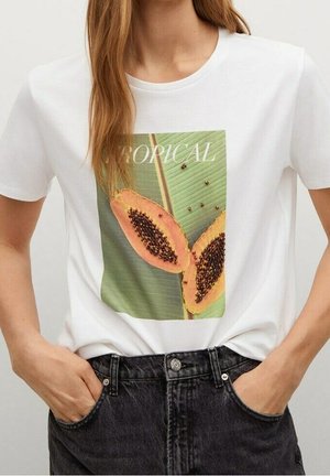 Femme portant une chemise blanche avec un graphisme de deux papayes coupées en deux et une feuille verte, avec le texte "TROPICAL" au-dessus.