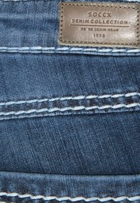 Denim w ciemnoniebieskim odcieniu o gładkiej fakturze. Posiada białe przeszycia oraz skórzaną łatę w kolorze brązowym oznaczoną napisem "SOCCX Denim Collection."