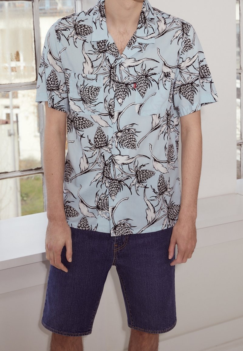 Camicia a maniche corte azzurro chiaro con motivi floreali e di piante neri, abbinata a pantaloncini di denim scuro. Design semplice, tasca sul petto visibile.