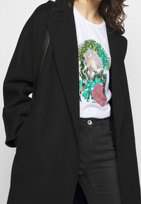 Black wool coat with a loose fit, worn over a white T-shirt featuring a colorful, sequin-embellished plant graphic, paired with black jeans.