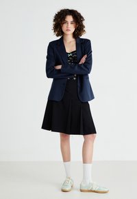 Námořnický blazer přes květinový top, kombinovaný s černou skládací sukní a mátovými teniskami. Viditelné bílé ponožky. Minimalistický a moderní design.
