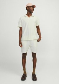 Set di polo e pantaloni corti in crema chiaro, misto cotone. Scarpe marroni con dettagli a fiocco e calze color sabbia. Tessuto morbido, vestibilità rilassata.