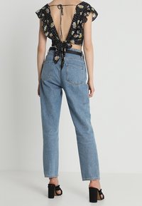 Femme portant un jean taille haute bleu clair, un crop top noir à motif floral avec dos ouvert noué par un nœud, et des sandales à talons larges noires, debout de dos.