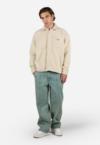 Beige fleece skjortejakke med krave, to brystlommer og frontknapper. Teal baggy jeans og hvide sneakers fuldender outfittet.