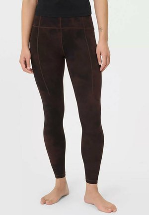 Persoon draagt donkerbruine legging met zichtbare stikseldetails, staat op blote voeten tegen een effen witte achtergrond.