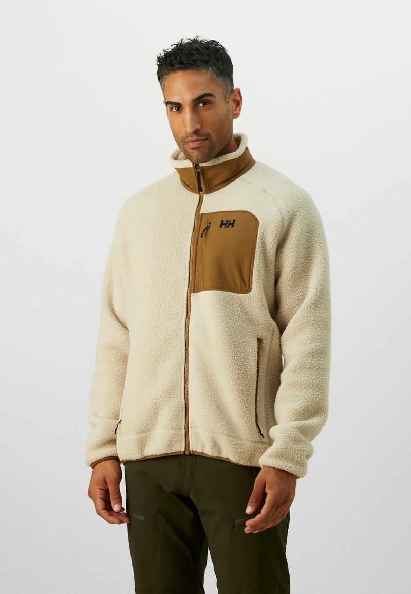Chaqueta de forro polar en crema con un bolsillo de pecho con cremallera en color marrón. Presenta un cuello alto, cierre frontal con cremallera y puños elásticos. Textura duradera y suave.