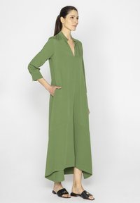 Zielona maxi sukienka z kołnierzykiem, rękawami o długości 3/4, bocznymi kieszeniami i asymetrycznym dołem. Noszona z czarnymi płaskimi sandałami.