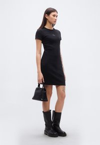 Μαύρο φόρεμα με κοντά μανίκια και εφαρμοστή σιλουέτα, με λεία υφή υφάσματος και κεντημένο λογότυπο. Συνδυασμένο με μικρή μαύρη τσάντα και μπότες μέχρι το γόνατο.