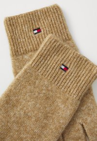 Guantes de color marrón tejido con puños acanalados, elaborados a partir de una tela texturizada. Pequeño logo en rojo, blanco y azul cerca de la muñeca.