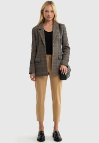 Raibais žakets brūnā, pelēkā un oranžā krāsā, ar vienu pogu un divām pogām, kombinēts ar bežām biksēm un spīdīgiem melniem mokasīniem.