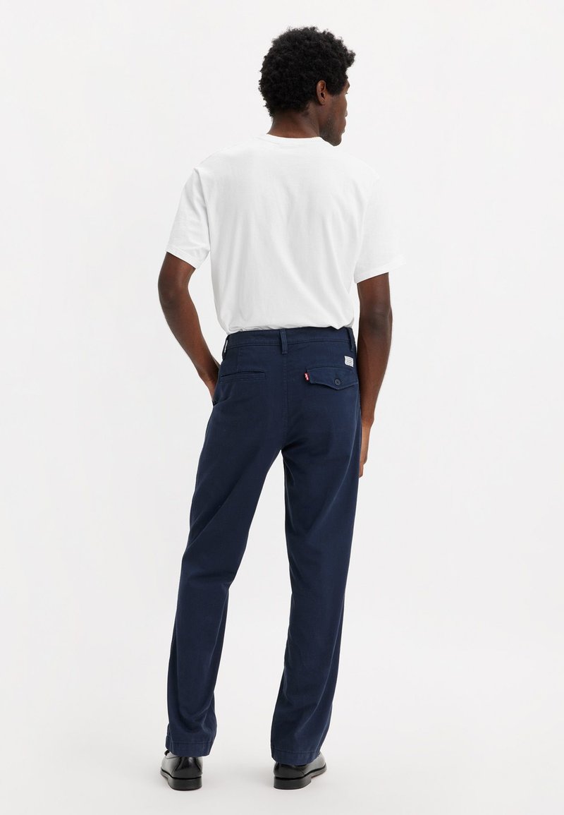 Pantalones azul marino con un diseño de pierna recta, bolsillo trasero abotonado y trabillas para cinturón, combinados con una camiseta blanca lisa.