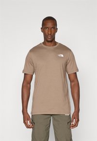 Tricou scurt din bumbac maro, cu mânecă scurtă, având un logo alb pe pieptul stâng, asociat cu pantaloni cargo verzi, evidențiind un stil lejer.