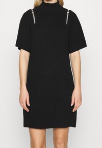 Robe en maille noire à manches courtes, col montant et coupe ample. Présente des accents de quincaillerie argentée sur les épaules et une poche avant.