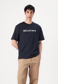 Tengerész kék t-shirt, kerek nyakkal és rövid ujjakkal, a mellkasán jól látható fehér "BELSTAFF" logóval. Bézs nadrággal viselve.