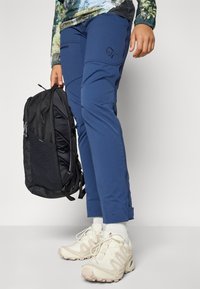 Plave vanjske hlače s prilagođenim dizajnom, s patentnim zatvaračima na džepovima i izvezenim logotipom, u kombinaciji s crnim ruksakom i bež tenisicama.