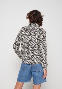Maglietta nera con motivo floreale bianco, maniche lunghe, colletto button-down, indossata con pantaloni corti in denim blu con bordi sfrangiati e un'etichetta sulla tasca posteriore.