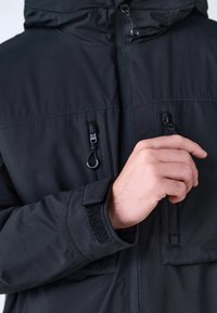 Mano ajustando la cremallera del pecho en una chaqueta negra con una correa de Velcro visible en la manga y la capucha en el cuello.