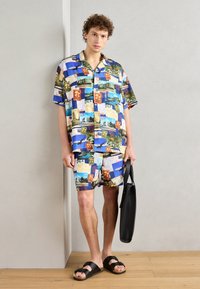 Kortærmet blå skjorte og matchende shorts præsenterer en collage af postkort med forskellige farver og billeder. Sort bæretaske og sandaler.