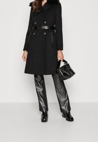 Vrouw in een zwart gebelt coat met zilveren knopen, metalen broek, zwarte hoge hakken, en houdt een zwarte leren handtas vast bij het hengsel.