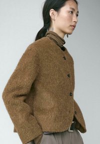 Brown fuzzy knit jacket with a boxy fit, featuring a round neckline, black buttons, and long sleeves. Layered over a brown turtleneck.