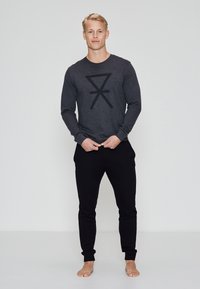 Camisa de manga larga de color gris oscuro con diseño gráfico a rayas negras, combinada con joggers negros ajustados. La tela parece suave; el modelo está descalzo.