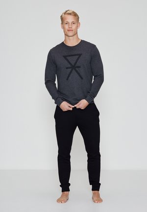 Camisa de manga larga de color gris oscuro con diseño gráfico a rayas negras, combinada con joggers negros ajustados. La tela parece suave; el modelo está descalzo.