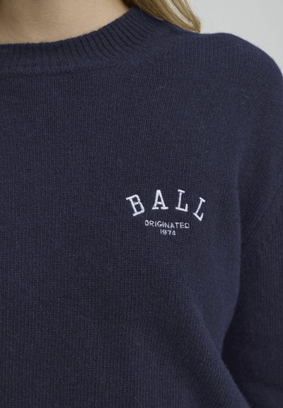 Marineblå strikkegenser med ribbestrikket krage og brodert hvit tekst "BALL" med "ORIGINATED 1974" under. Myk tekstur, avslappet passform.