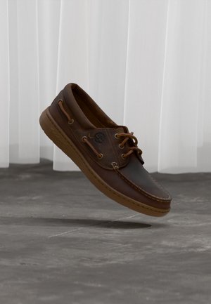 Hnědá kožená bota typu boat shoe se šňůrkami, logo Timberland na boku, vznášející se nad šedou dlažbou před bílými vertikálními žaluziemi.