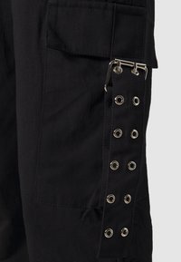 Pantalon en tissu noir avec grande poche latérale et sangle décorative dotée d'œillets en métal argenté et de détails à boucle.