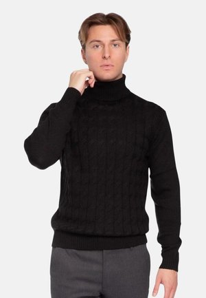 Giovane uomo che sistema un maglione nero a collo alto lavorato a trecce, in piedi davanti a uno sfondo chiaro e semplice, indossando pantaloni grigi.