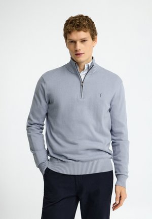 Giovane uomo che indossa un maglione grigio chiaro con zip al quarto sopra una camicia bianca con colletto e pantaloni scuri, in piedi con una mano in tasca.