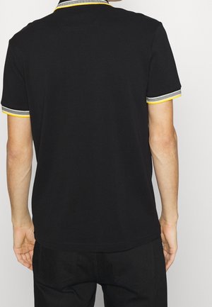 Mann trägt schwarzes kurzärmeliges Polo mit gelb-grau gestreiftem Kragen und Ärmelsäumen, steht mit dem Rücken zum Betrachter vor einem schlichten Hintergrund.