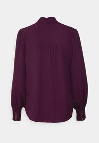 Blouse mauve foncé à manches longues avec col montant, empiècement plissé dans le dos et poignets boutonnés avec œillets dorés vue de dos.