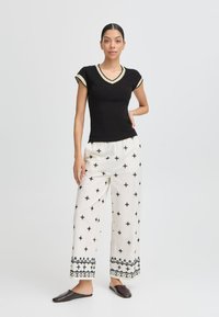 Tricou negru cu decolteu în V și mâneci scurte, asortat cu pantaloni albi largi, confeționați cu modele brodate negre și o margine decorativă. Pantofi plată maro.