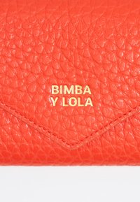 Izšūts oranžs ādas maks ar izteiktu zelta logo "BIMBA Y LOLA", piedāvājot trīsstūrveida vāku un kontrastējošiem dūrieniem.