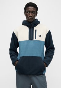 Vīrietis, kurš valkā krāsu bloku flīsa kapuci džemperi ar tumši ziliem, gaiši zilajiem un krēmkrāsas laukumiem, rokām priekšējā kabatā, skatās uz sāniem.