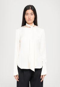 Bluză de univor deschis, cu cravată la gât, m�âneci lungi și o croială ușor lejeră. Țesătura pare netedă, cu o strălucire subtilă. Detalii cu manșete butonate.