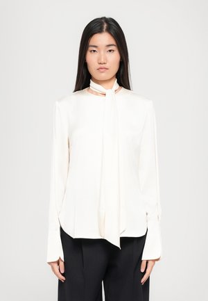 Chemisier ivoire clair avec un nœud au cou, manches longues et une coupe légèrement ample. Le tissu semble lisse avec un léger éclat. Détails des poignets boutonnés.